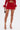 A person wears the FIERCE MESH MINI SKIRT in red, a matching long-sleeve top with feathered cuffs, gold strappy heels, and carries a small brown patterned handbag against a plain white background.