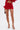 A person wearing the FIERCE MESH MINI SKIRT, a red feather-trimmed top, and gold strappy heels stands against a white background, holding a small brown purse.