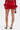 A person poses on a white background wearing the FIERCE MESH MINI SKIRT in red with a matching top featuring feathered cuffs, high-heeled ankle-strap sandals, and holding a small brown handbag.