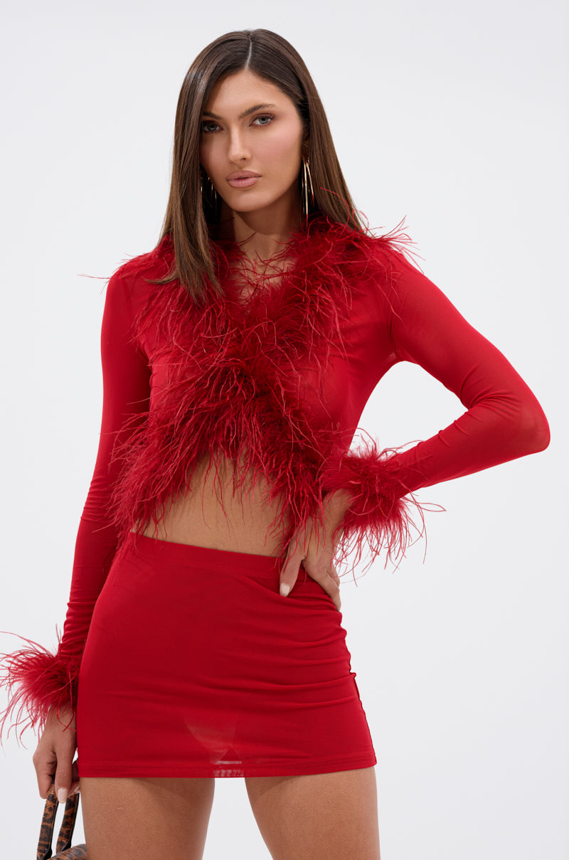 A woman stands against a plain light background with one hand on her hip, wearing the FIERCE FEATHER BLOUSE—a red, sheer mesh top with feather trim—paired with a matching red mini skirt.