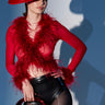 A woman poses in a dramatic red wide-brimmed hat, the FIERCE FEATHER BLOUSE with feather trim and sheer mesh, black high-waisted shorts, and holds a shiny red sculptural accessory against a gray background.