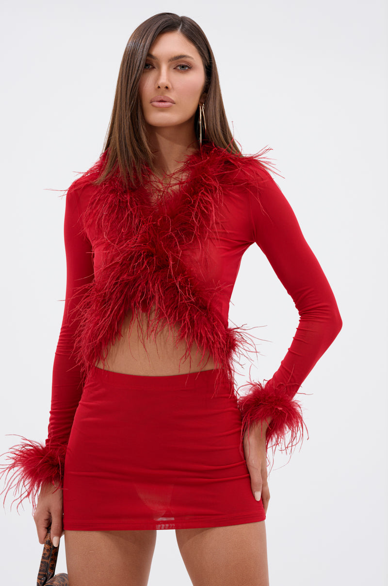 A woman with straight brown hair and a neutral expression stands confidently against a plain light background, wearing the FIERCE FEATHER BLOUSE—a red sheer mini dress with long sleeves and bold red feather trim.