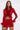 A woman with straight brown hair and a neutral expression stands confidently against a plain light background, wearing the FIERCE FEATHER BLOUSE—a red sheer mini dress with long sleeves and bold red feather trim.