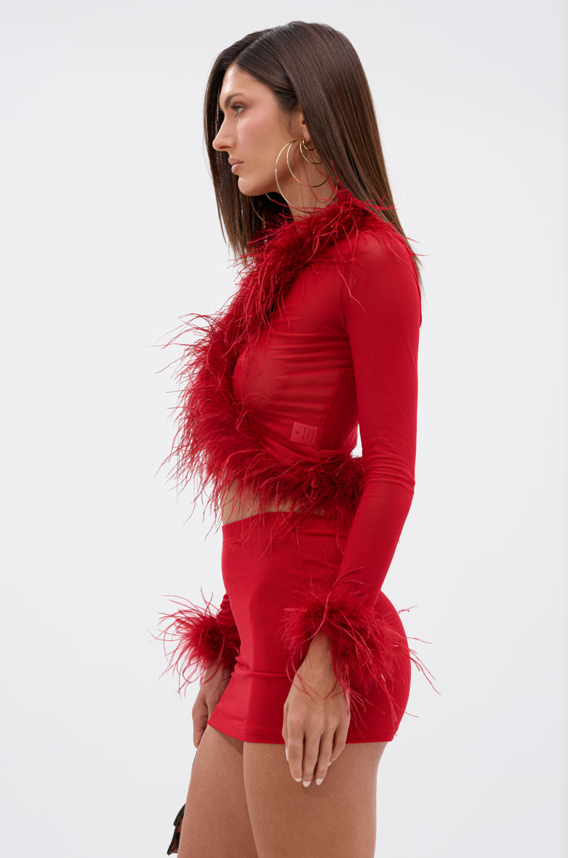 A woman stands in profile wearing the FIERCE FEATHER BLOUSE—a fitted, long-sleeve red crop top with a matching mini skirt, both detailed with red feather trim on the neckline, sleeves, and hem—set against a plain light background.