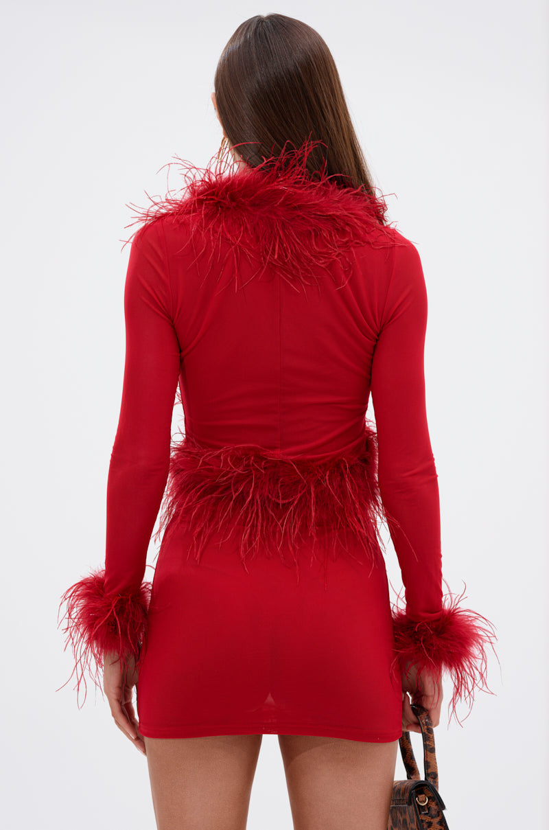 A woman with long brown hair, seen from behind, wears the FIERCE FEATHER BLOUSE—a fitted red blouse with feather trim and sheer mesh top—while holding a small purse. The background is plain white.