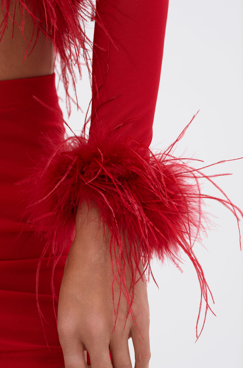 Wearing the FIERCE FEATHER BLOUSE, a bold red AKIRA Label long-sleeve mesh top with dramatic feathered cuffs and paired with a skirt, stands out vividly against a plain white background.