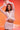 A woman with long dark hair and a serious expression raises her arms above her head while posing confidently in the ICY HOT FEATHER BLOUSE against a vibrant pink and orange gradient background.