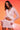 A woman poses confidently in the ICY HOT FEATHER BLOUSE, a light pink, feather-trimmed, long-sleeve crop top with a matching mini skirt, set against a warm orange-pink gradient background.