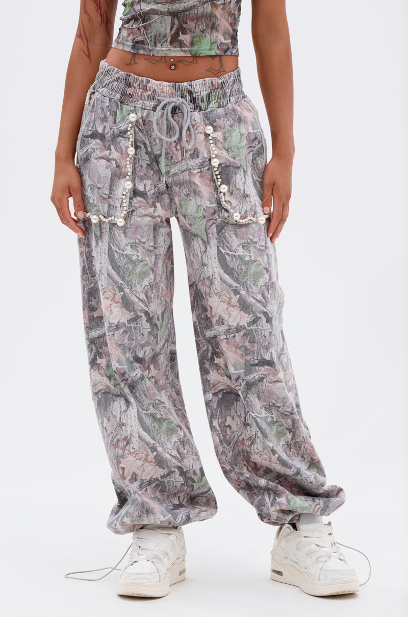 A person models the MEET ME HALFWAY WIDE LEG PANT—loose, camo print cargo pants with beaded and shell pocket details—paired with a matching top and white sneakers, standing against a plain white background. 0