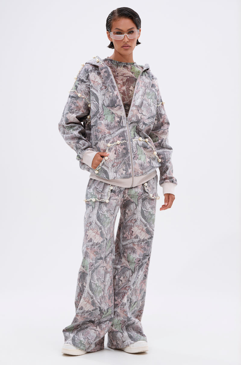 A person confidently poses against a white background wearing the MEET ME HALFWAY WIDE LEG PANT in a loose camo print with light accents and side tassels, paired with a matching jacket, sunglasses, and white shoes. 6