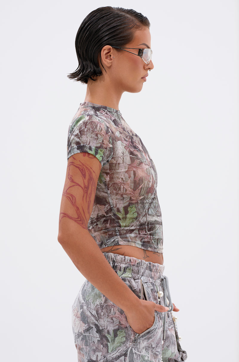 A person with slicked-back hair and sunglasses stands in profile, wearing the MEET ME HALFWAY PRINTED MESH TEE and matching camo shorts with a cropped fit. They have a tattoo on their arm and hands in pockets, against a plain light background.