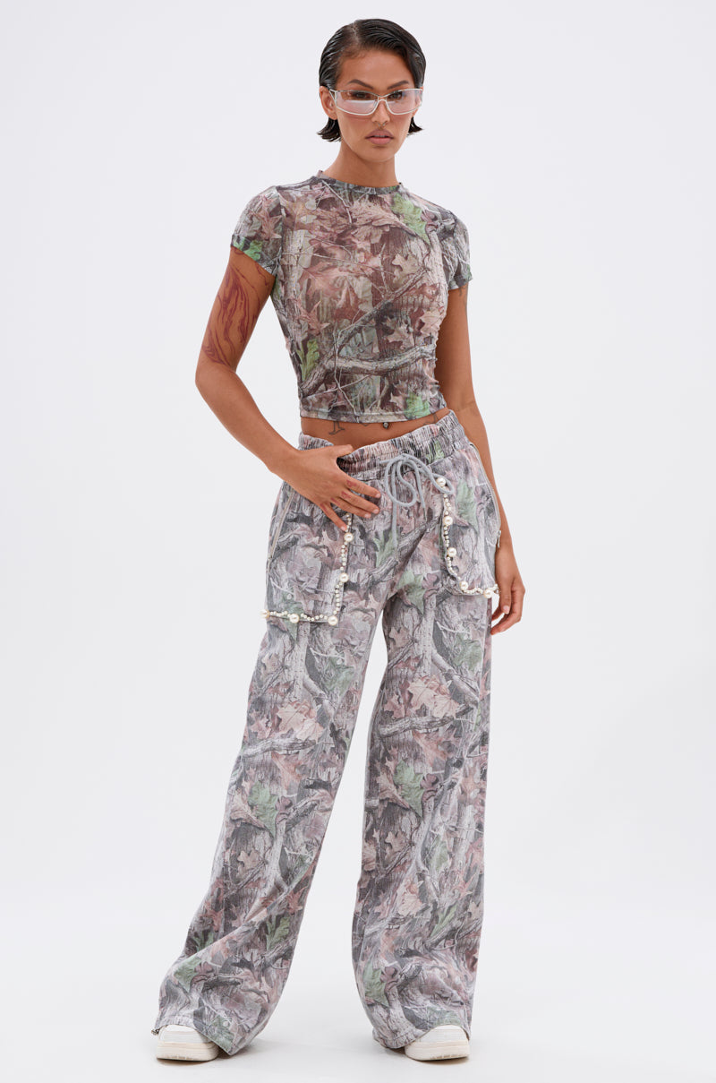 A person stands confidently in the MEET ME HALFWAY PRINTED MESH TEE, a cropped short-sleeve top with a muted abstract camo print, paired with wide-leg pants, clear glasses, and white sneakers against a plain light background.