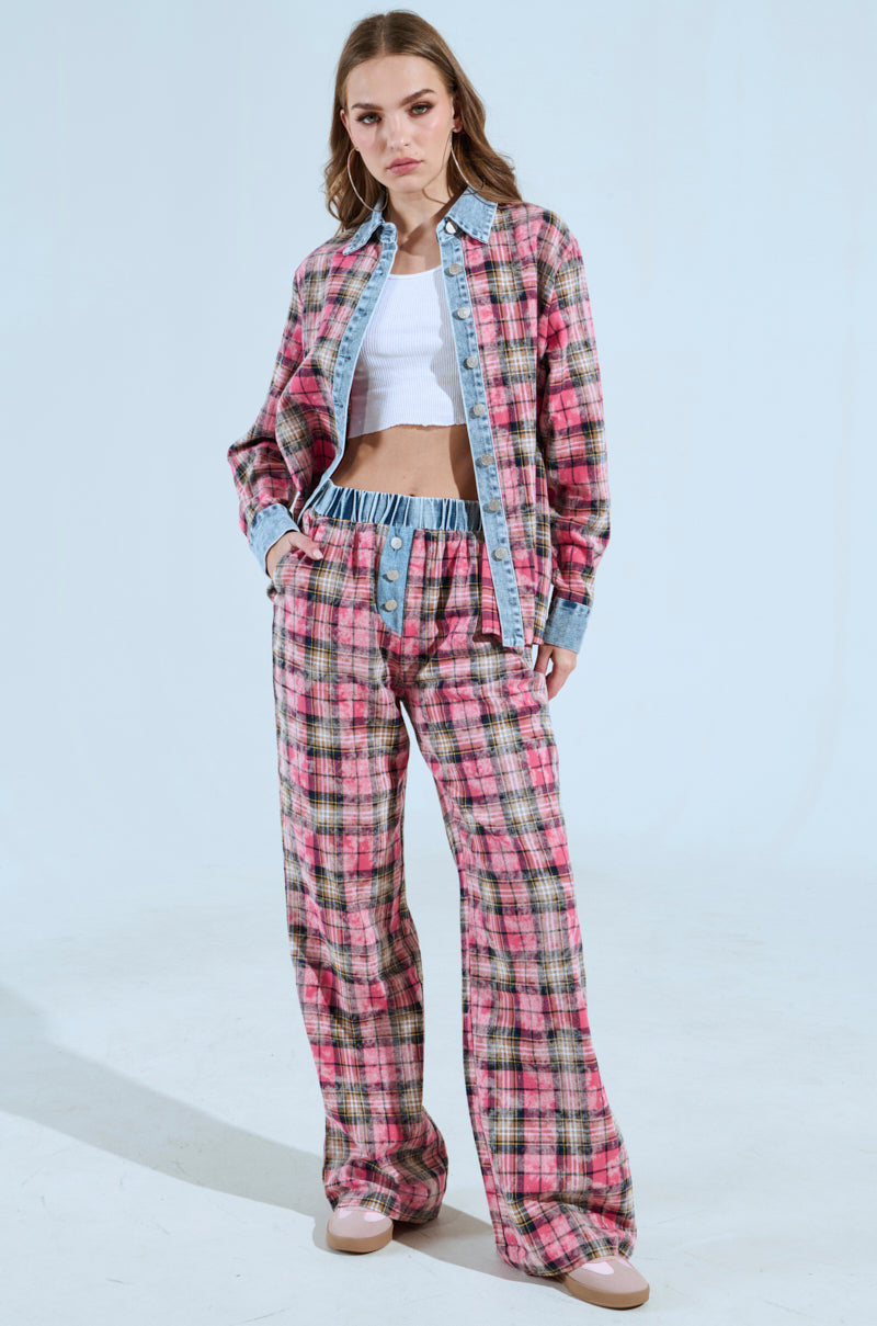 A person is dressed in the "THE ONE PINK PLAID WIDE LEG PANT" paired with a matching pink and gray plaid jacket over a white crop top. The outfit, featuring light denim accents in a high-rise silhouette, is set against a light blue background while they maintain a neutral expression.