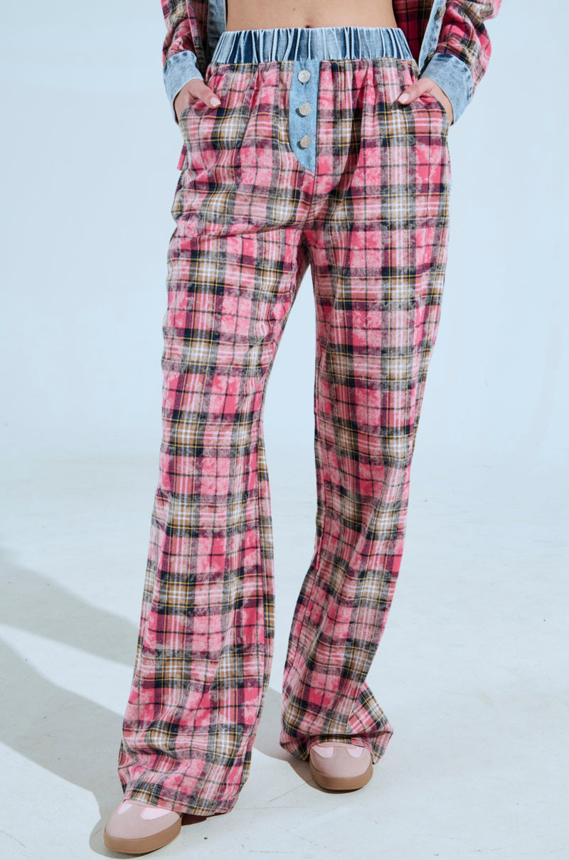 A person models THE ONE PINK PLAID WIDE LEG PANT, featuring a custom button design and denim waistband with a high-rise silhouette, paired stylishly with a plaid shirt and light pink shoes against a minimalist backdrop.