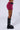 A person stands sideways, modeling the WHATEVER SHE WANTS MINI SKIRT in a vibrant magenta hue, paired with tall, black chunky boots adorned with multiple silver buckles and straps. The skirt's sleek design is enhanced by black belt details that add an edgy sophistication against the plain white background.