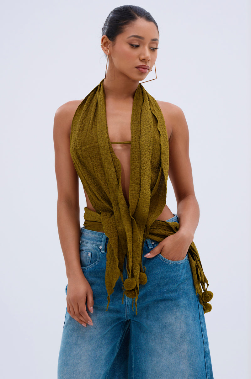 Wearing the ISLAND DREAMS WRAP HALTER TOP in crinkled olive knit with fringe and pom-pom details, a woman pairs it with high-waisted blue jeans, her hair tied back as she poses with one hand in her pocket.