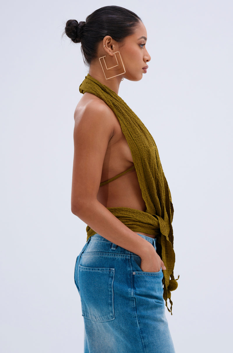 A woman in profile wears the ISLAND DREAMS WRAP HALTER TOP, a convertible crinkled olive knit, paired with blue jeans. Her hair is in a neat bun, and she wears large geometric gold earrings against a plain light background.