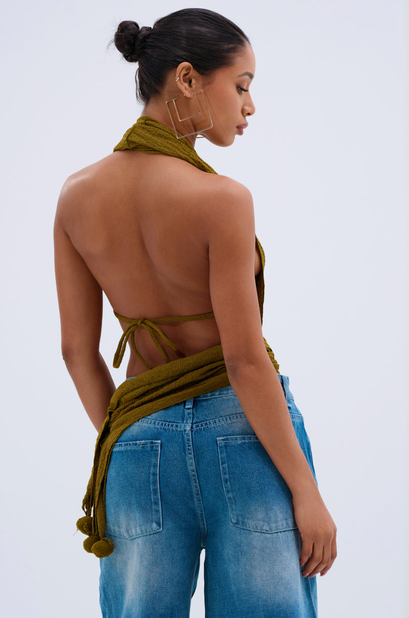 A woman with her hair in a bun wears the ISLAND DREAMS WRAP HALTER TOP in crinkled olive knit and blue high-waisted jeans, facing away to highlight its open back, decorative tassels, and large geometric earrings.