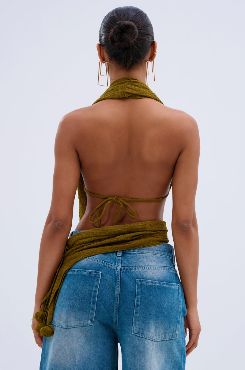 A woman with her hair in a bun is seen from behind, wearing the ISLAND DREAMS WRAP HALTER TOP in crinkled olive knit and loose blue jeans, her top partly wrapped at the waist and accessorized with bold geometric gold earrings.