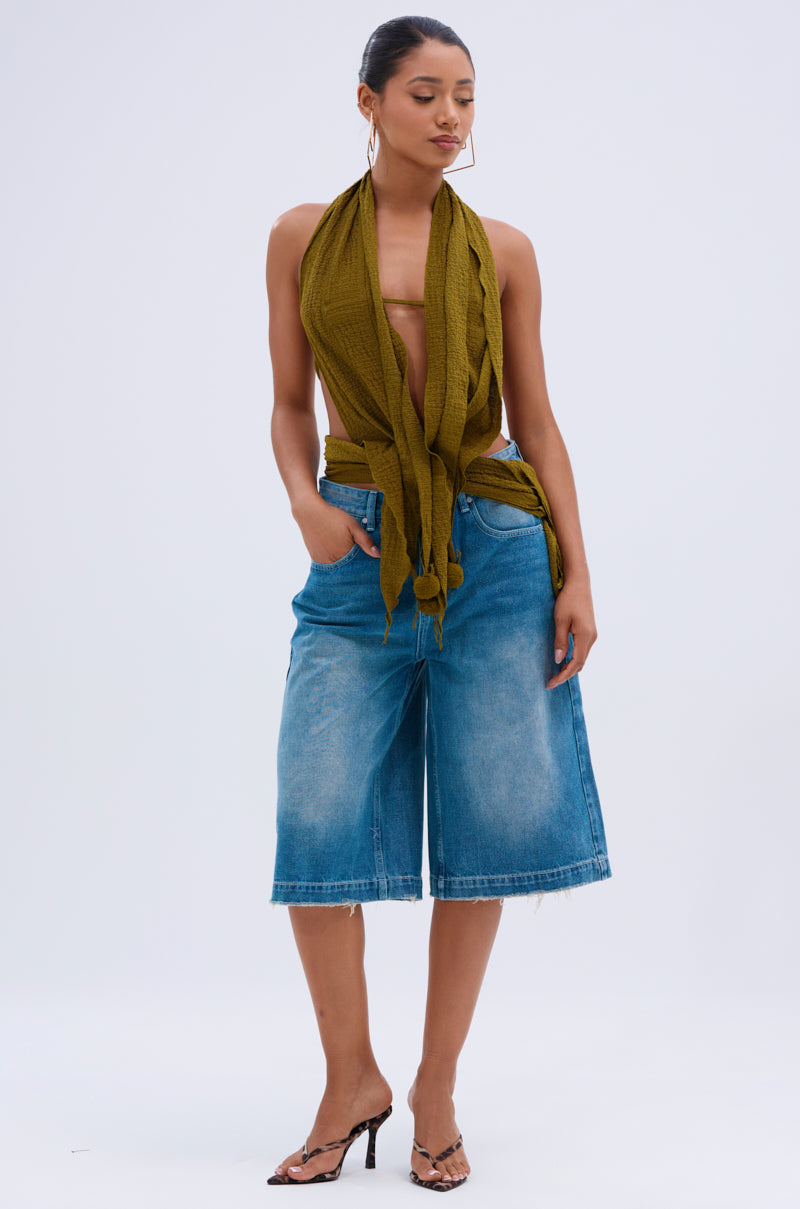 A woman wearing the ISLAND DREAMS WRAP HALTER TOP in crinkled olive knit, blue knee-length denim shorts, and heeled sandals stands with her hands in her pockets against a plain white background.