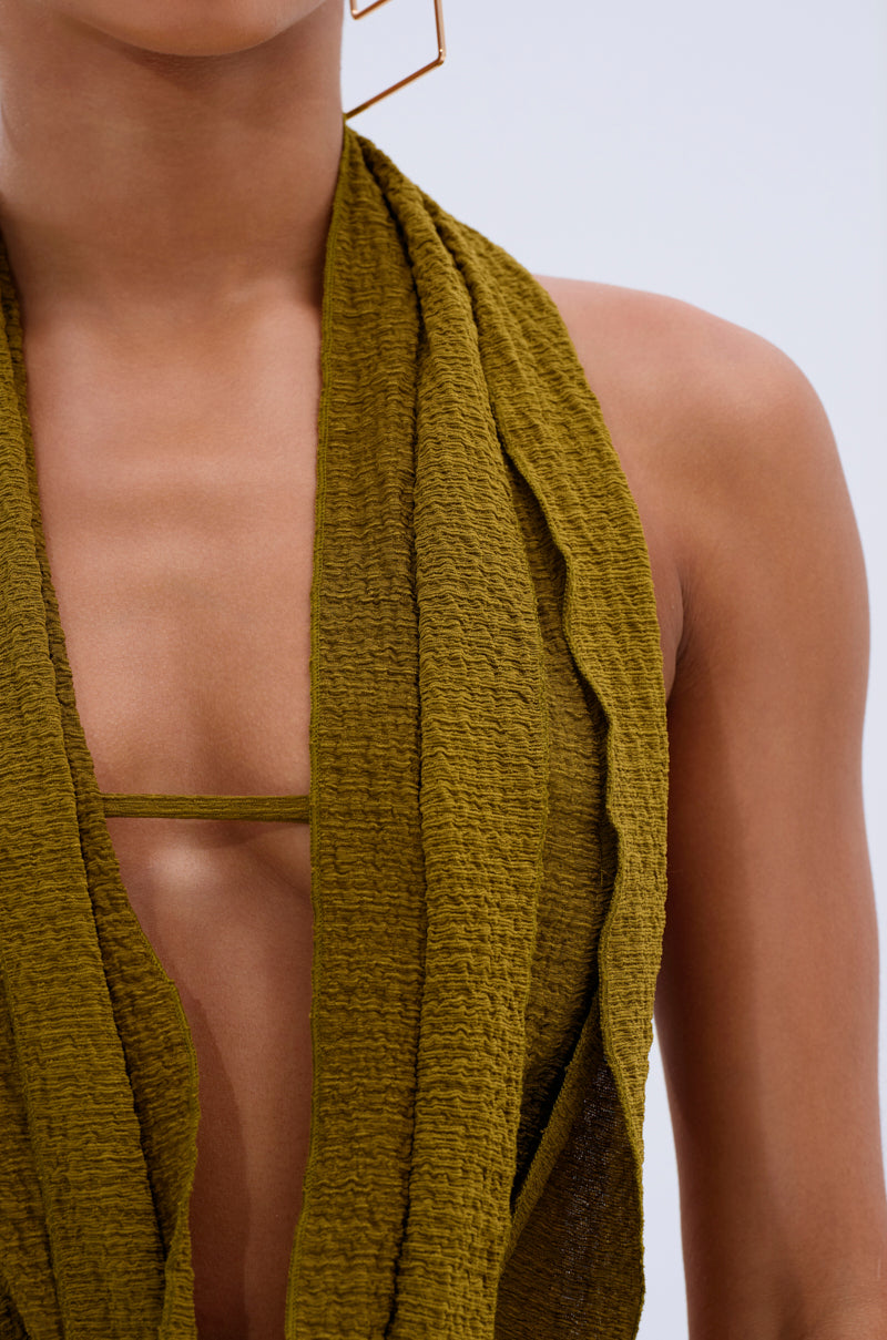 A close-up shows someone wearing the ISLAND DREAMS WRAP HALTER TOP in crinkled olive knit, featuring a deep neckline and thin straps; this versatile, convertible piece pops against a plain, light background.