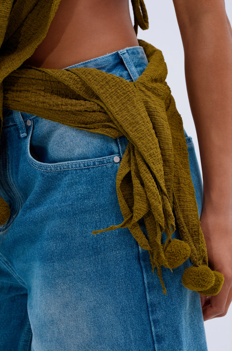 Close-up of a person in blue jeans and the ISLAND DREAMS WRAP HALTER TOP, a crinkled olive knit halter with pom-pom accents, tied at the waist to show their midriff, hand resting by their side.