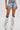 Wearing the DIVALICIOUS DENIM SHORT—light blue with floral embroidery, pearls, and fringe—styled Western with silver metallic cowboy boots with long white fringe. Shot against a plain white background.