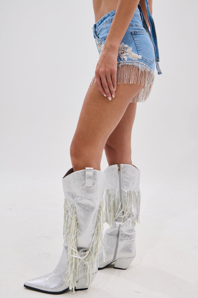 A person wears DIVALICIOUS DENIM SHORTS with Western-style rhinestone embellishments and shiny silver fringe cowboy boots, standing against a plain white background.