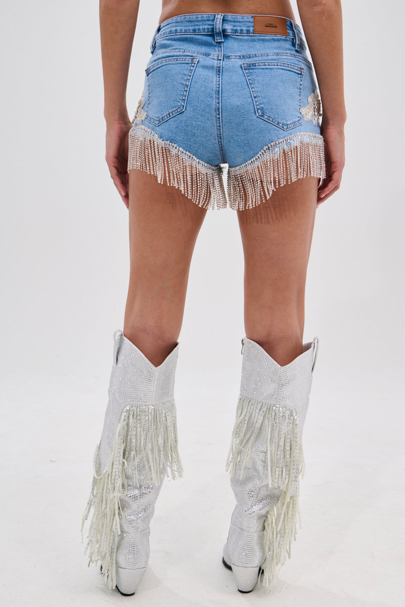A person faces away, wearing DIVALICIOUS DENIM SHORTS with pearl fringe trim and sparkly white Western-style fringe cowboy boots against a plain white background.