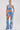 A woman faces forward in Western style, wearing the DIVALICIOUS DENIM SHORT paired with a halter crop top and chaps-style jeans embellished with lace appliqué and fringe, plus slicked-back hair, hoop earrings, and white pointed boots.