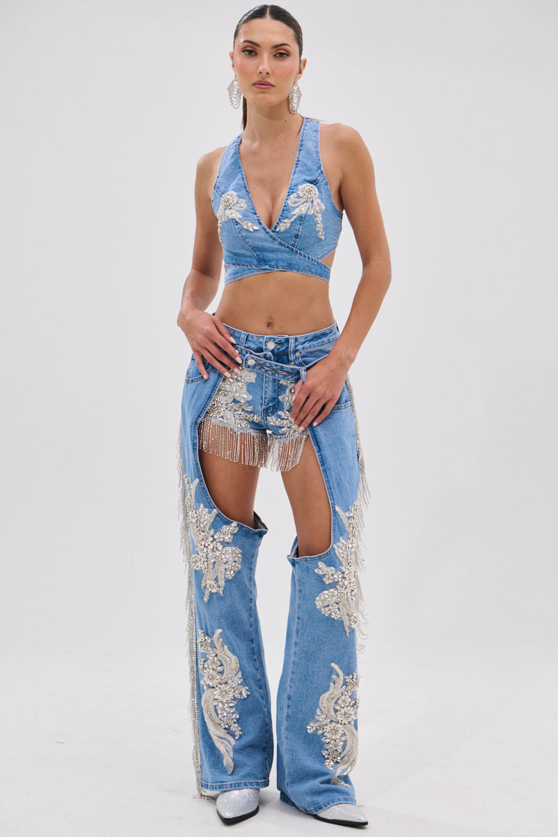 A woman faces forward in the DIVALICIOUS DENIM CHAPS IN LIGHT BLUE with silver embroidery and rhinestone chain link fringe, styled with a cropped halter top, high-waisted shorts, white boots, slicked-back hair, and large earrings.