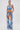 A woman faces forward in the DIVALICIOUS DENIM CHAPS IN LIGHT BLUE with silver embroidery and rhinestone chain link fringe, styled with a cropped halter top, high-waisted shorts, white boots, slicked-back hair, and large earrings.
