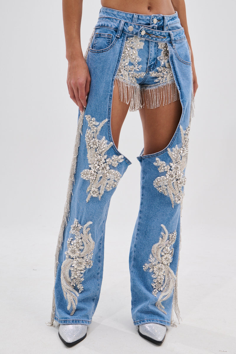 Someone wears DIVALICIOUS DENIM CHAPS IN LIGHT BLUE with large cutouts exposing beaded fringe shorts and rhinestone chain link fringe. The chaps have silver embroidery, pearl-like accents, and are styled with pointed silver shoes.