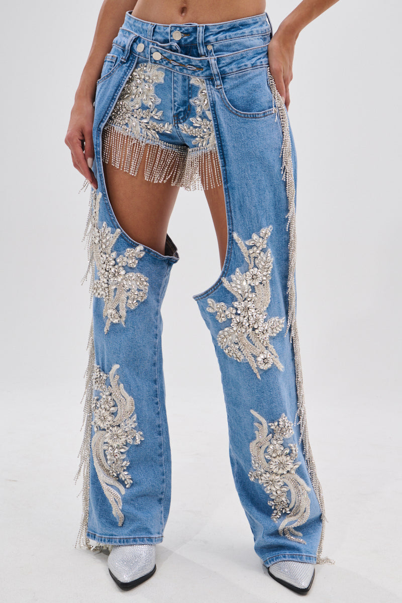 A person wears DIVALICIOUS DENIM CHAPS IN LIGHT BLUE—jeans with one leg cut out to reveal beaded fringe shorts, rhinestone chain link fringe, silver embroidery, and sparkling bead details.