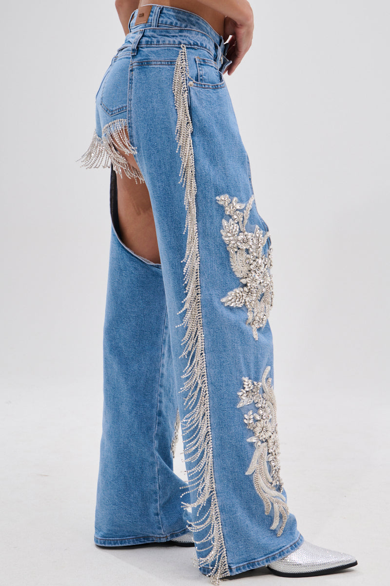 Wearing DIVALICIOUS DENIM CHAPS IN LIGHT BLUE, featuring intricate silver embroidery, rhinestone chain link fringe on sides and back, large outer thigh cutouts, and paired with silver pointed boots.