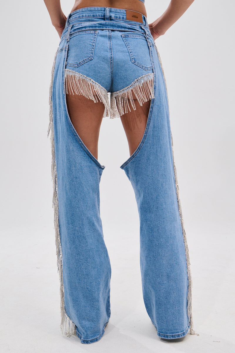 A person is shown from the back against a white background, wearing DIVALICIOUS DENIM CHAPS IN LIGHT BLUE with open sides and rhinestone chain link fringe over shorts.
