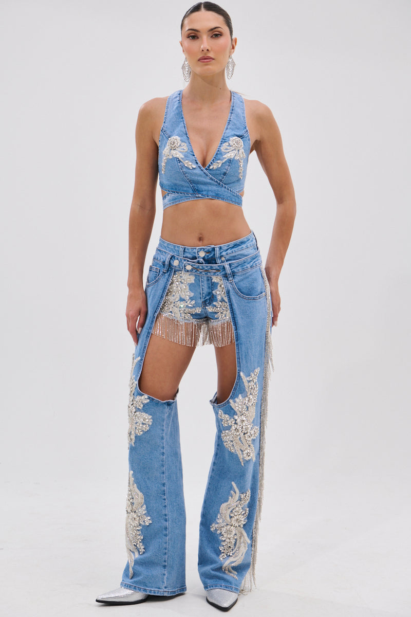A woman faces forward in the DIVALICIOUS DENIM CHAPS IN LIGHT BLUE— a sleeveless crop top, high-waisted shorts, and chaps-style pants with open fronts, beaded fringe, and sparkling rhinestone chain link fringe. She finishes the look with pointed white boots.