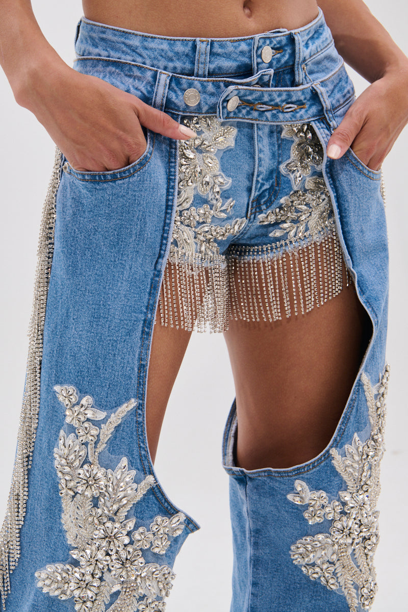 A person wears DIVALICIOUS DENIM CHAPS IN LIGHT BLUE featuring intricate silver floral patches, open at the front to reveal matching denim shorts accented with sparkling rhinestone chain link fringe.