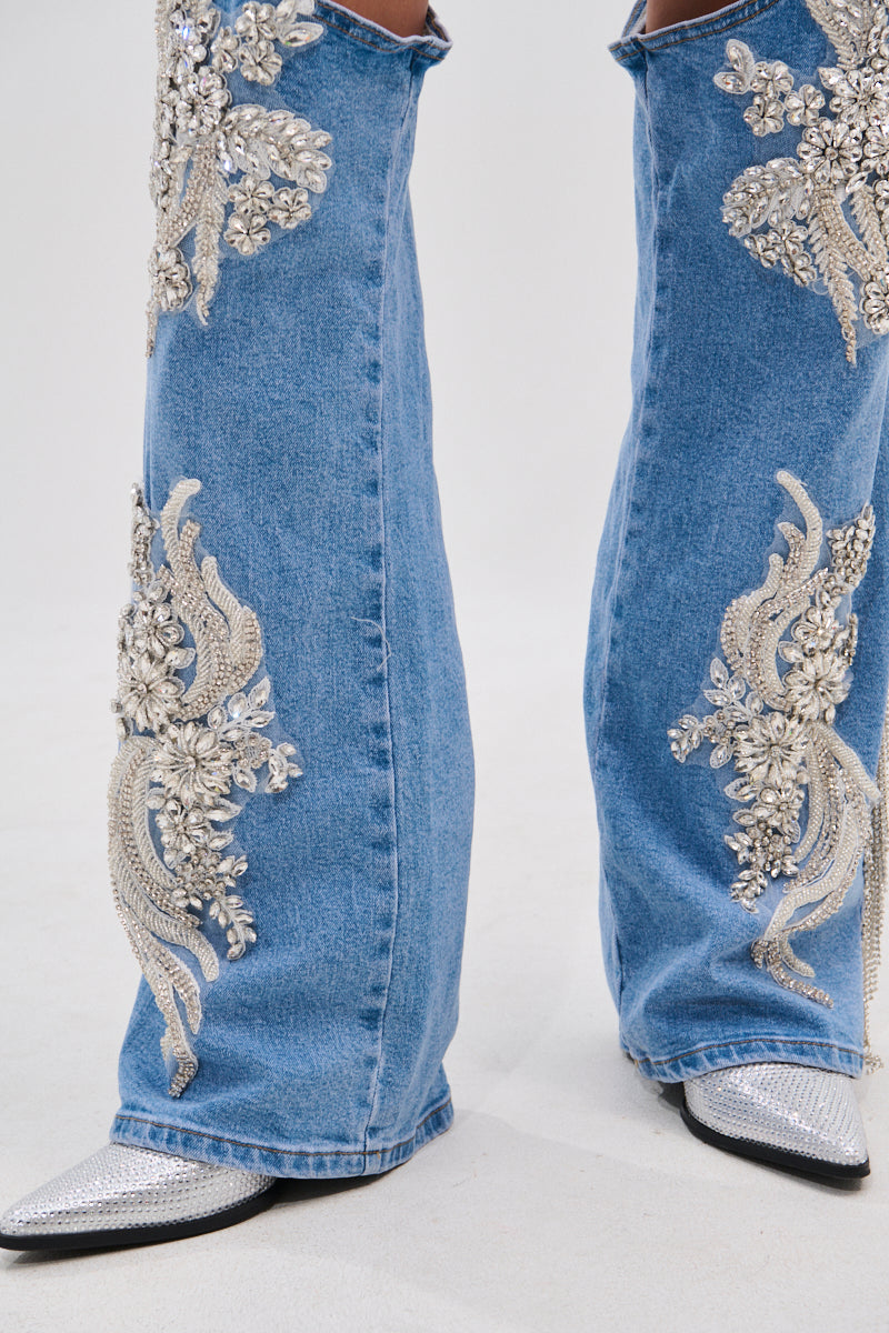 A close-up of legs wearing the DIVALICIOUS DENIM CHAPS IN LIGHT BLUE, featuring flared blue denim with silver bead and sequin floral designs, floral patches, pointed silver shoes, and sparkling rhinestone chain link fringe.