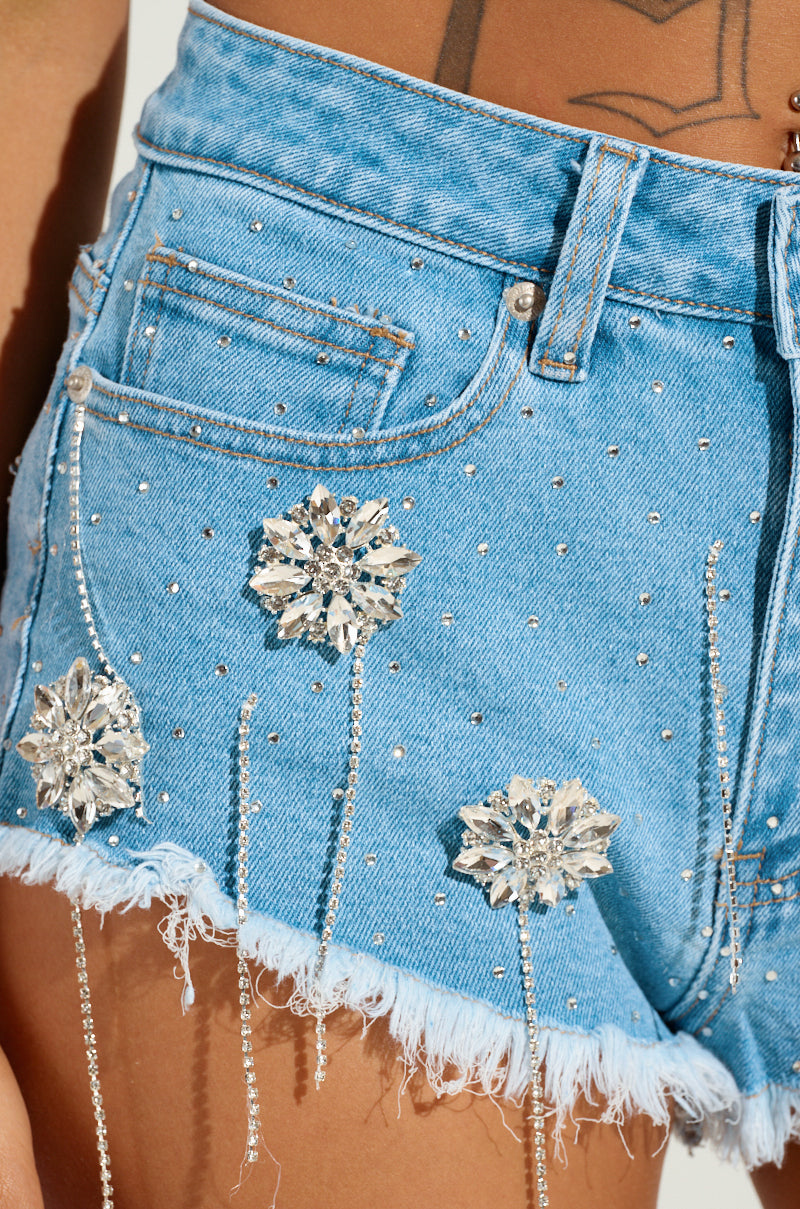 A close-up of a person's waist features the DIVALICIOUS DENIM MINI SHORT, adorned with rhinestone detailing and striking crystal floral embellishments. The frayed hems contribute an edgy touch, while a tattoo subtly reveals itself, adding personal flair to the ensemble.