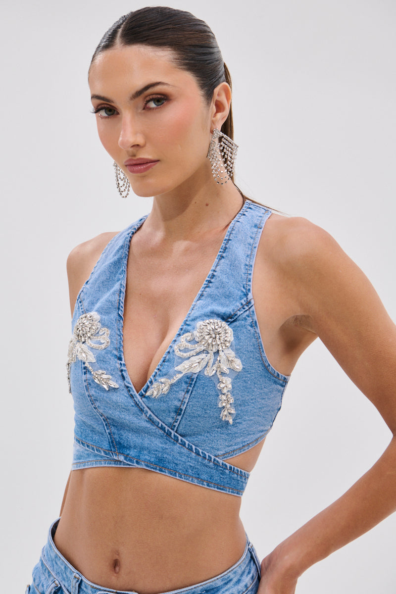 She wears the DIVALICIOUS DENIM BRA TOP IN LIGHT BLUE, a sleeveless deep V-neck style with rhinestone and silver floral details, paired with matching denim bottoms. Her long dark hair is pulled back, and she adds statement earrings for a western-glam finish.
