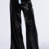 A person wearing the TRUE STORY SEQUIN PANT showcases a high-rise wide-leg silhouette adorned with shiny black sequins and complemented by a blue and white striped elasticized waistband. The pants are stylishly paired with black pointed-toe heels, all set against a pristine white background.