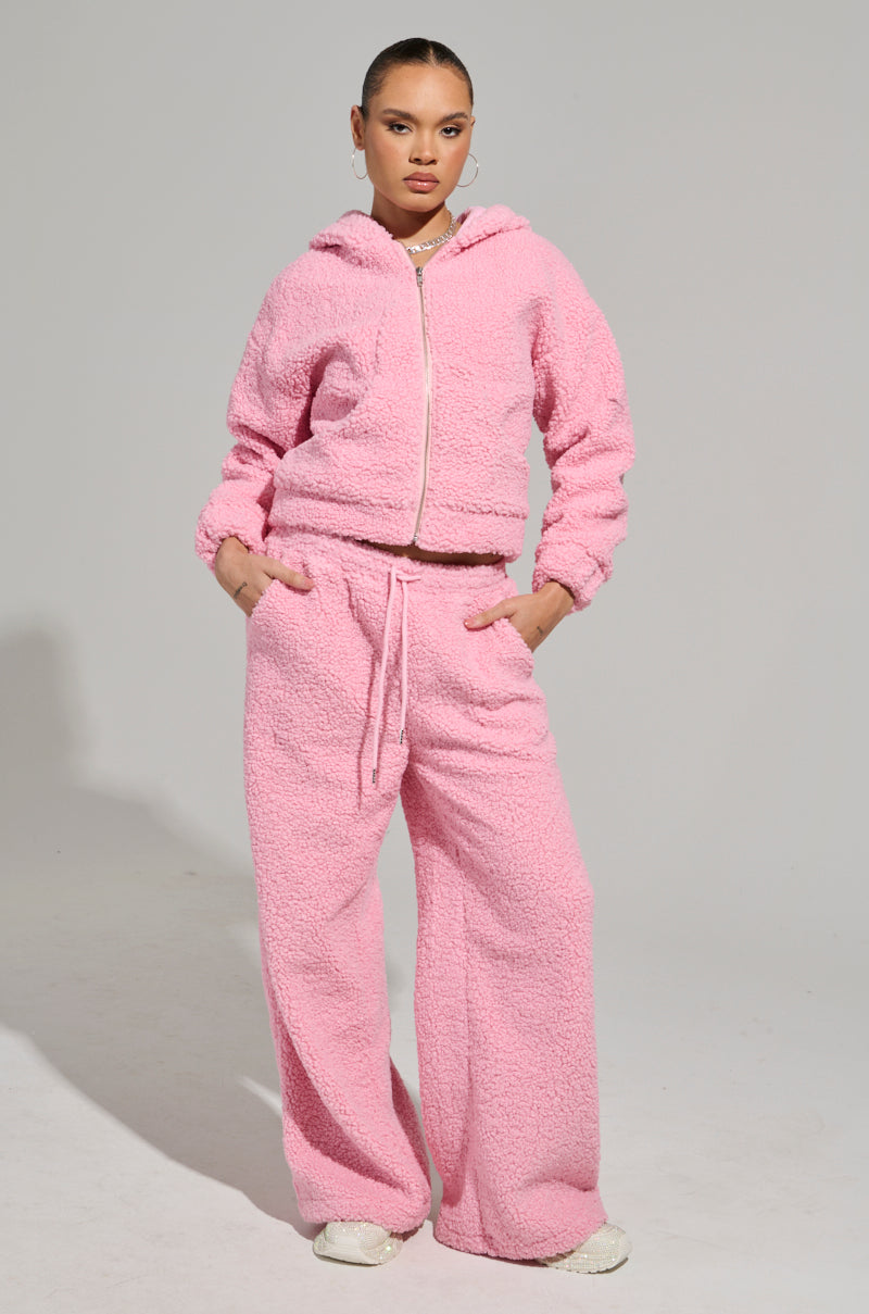 Dressed in a plush light pink hoodie, paired with the matching SOUR CANDY SHERPA JOGGER pants, an individual exudes confidence. Their hands rest in the pockets of this cozy ensemble as they complete the look with white sneakers against a minimalist light gray background, showcasing a perfect blend of casual charm.