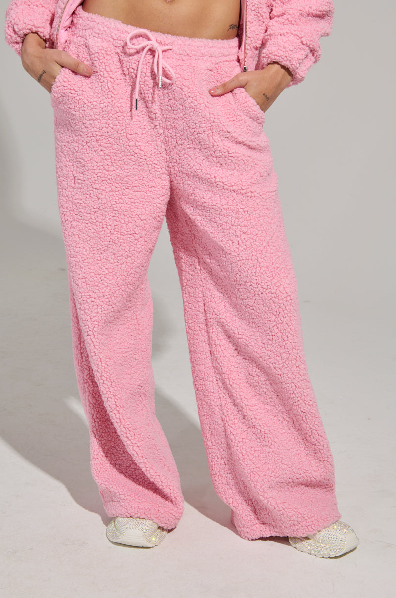 A person wearing comfortable, pink SOUR CANDY SHERPA JOGGERs with hands in pockets. These joggers feature a Sherpa fabrication and a drawstring waist, perfectly paired with white sneakers, standing against a plain background for the ultimate loungewear style.