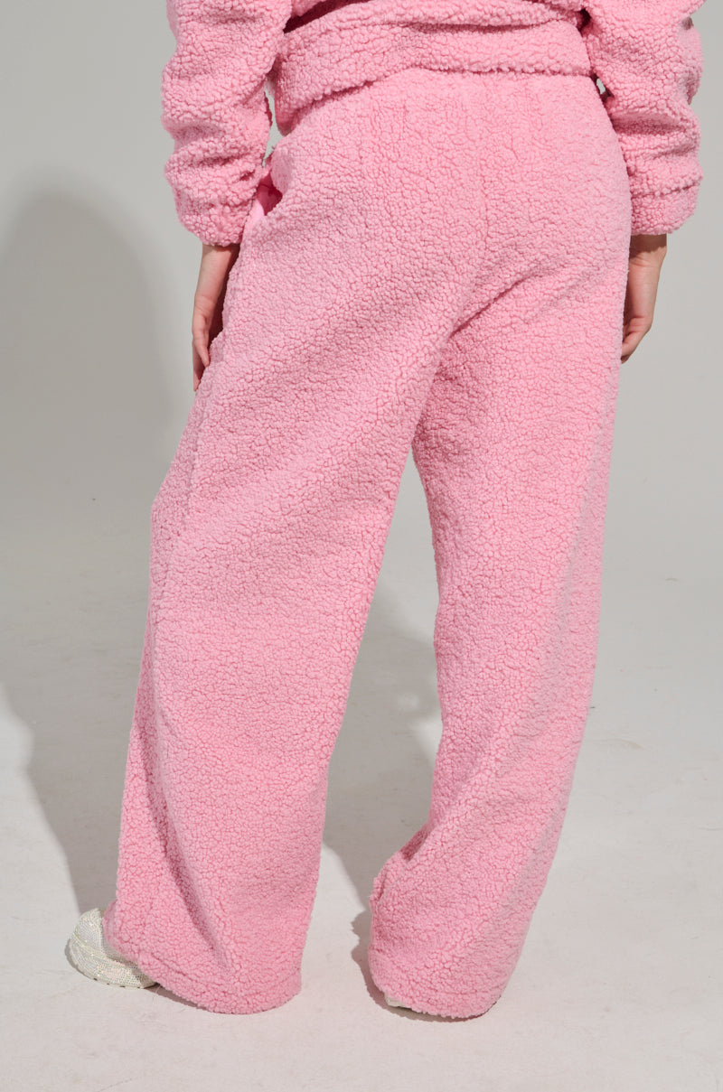 A person wearing the SOUR CANDY SHERPA JOGGER in pink, paired with a matching top, stands with their back to the camera. The sherpa fabric looks soft and inviting, capturing the spirit of loungewear. The background is plain and neutral.