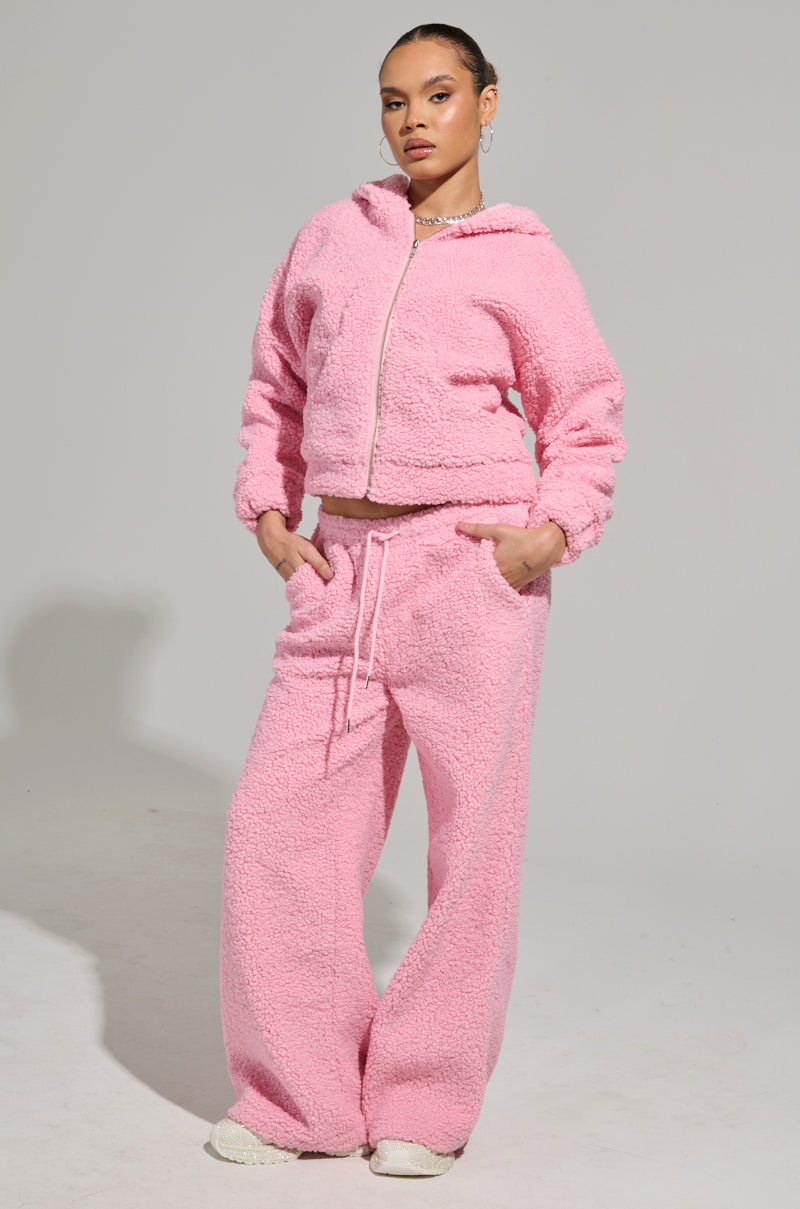 A person showcases the plush loungewear appeal of the SOUR CANDY SHERPA JOGGER tracksuit, complete with a hooded zip-up jacket and drawstring pants made from soft Sherpa fabric. The model poses confidently against a plain gray background.