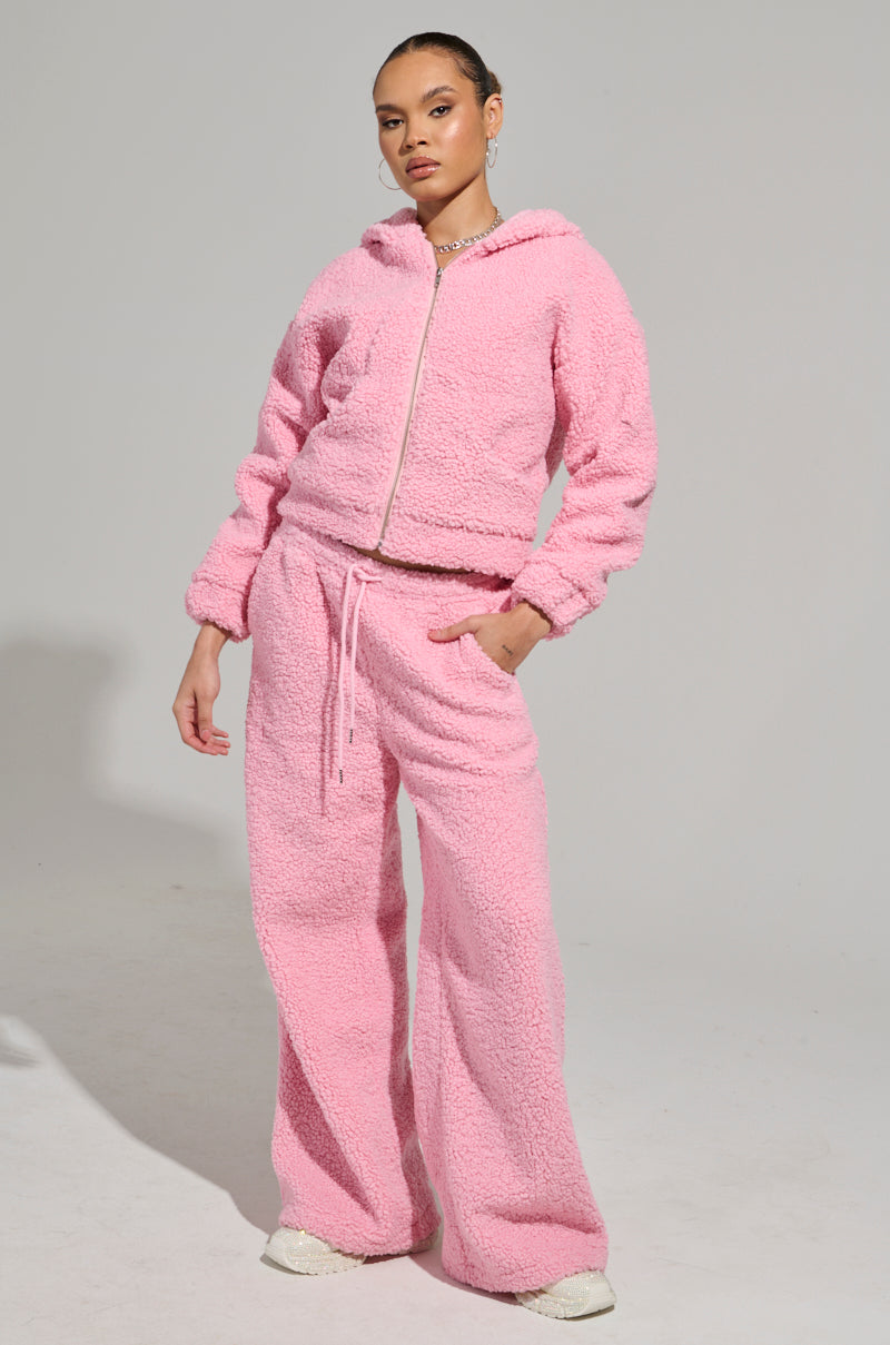 Dressed in a cozy pink tracksuit featuring wide-legged pants and a zip-up top, they stand stylishly against a light backdrop. The outfit evokes the vibe of the SOUR CANDY SHERPA JOGGER collection, complemented by white shoes and an aura of relaxed loungewear comfort.