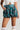 A person wearing a white top with laces paired with the LUCKY ME PLAID SHORT IN GREEN MULTI, a teal, high-waisted, plaid mini-skirt with a voluminous, bubble-like form. They have one hand in the skirt pocket and are also wearing thigh-high white lace-trimmed socks. This ensemble captures a bold plaid pattern perfect for the high-rise style trend.