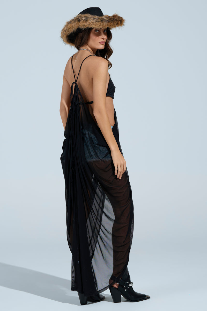 A woman embodies Coachella vibes in a festival-ready black ensemble, featuring the HEAVENLY THOUGHTS SLEEVELESS TUNIC TOP, sheer flowing cover, fur-trimmed hat, and loose waves. She looks back while facing sideways against a light blue backdrop, completing her look with black boots.