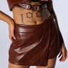 Person wearing a cropped dark brown faux leather top with button details and a matching high-waisted LOVE IN THIS CLUB SHORT from the AKIRA Label. A bold "1994" tattoo is visible on the lower abdomen and a detailed skull tattoo is on the thigh.